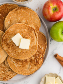 A top view of applesauce pancakes on a white plate with cinnamon and apples in the background.