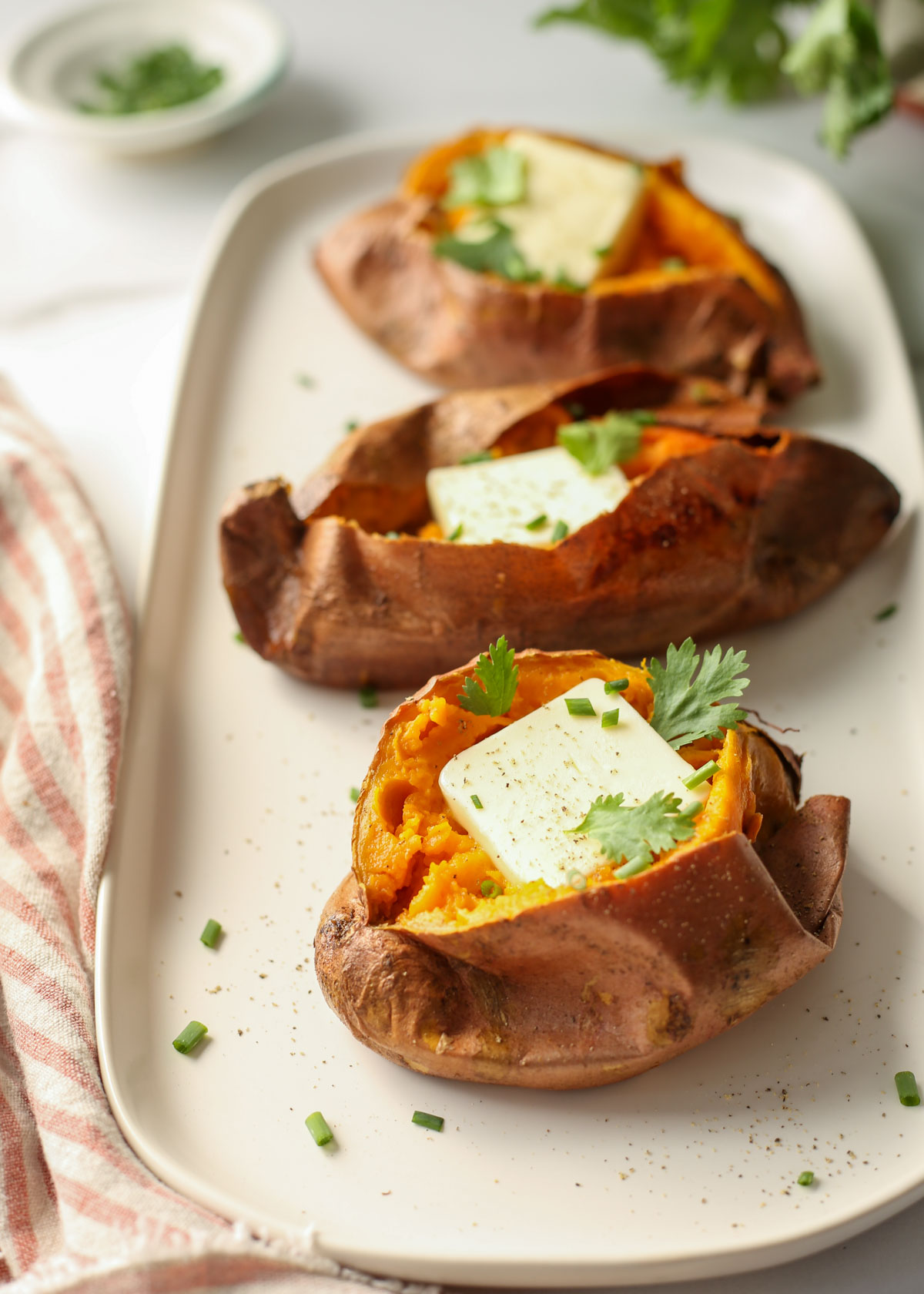 Three baked potatoes on a white platter with pats of butter, cilantro, and chives. 