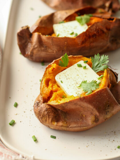 Three baked potatoes on a white platter with pats of butter, cilantro, and chives.