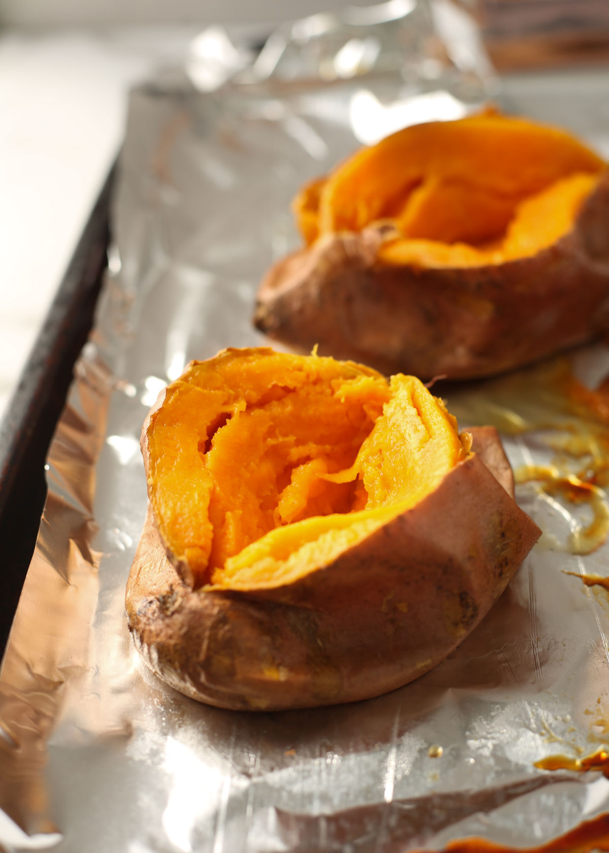 Two sweet potatoes on a foil-lined cookie sheet, ready to stuff with butter and other goodies.