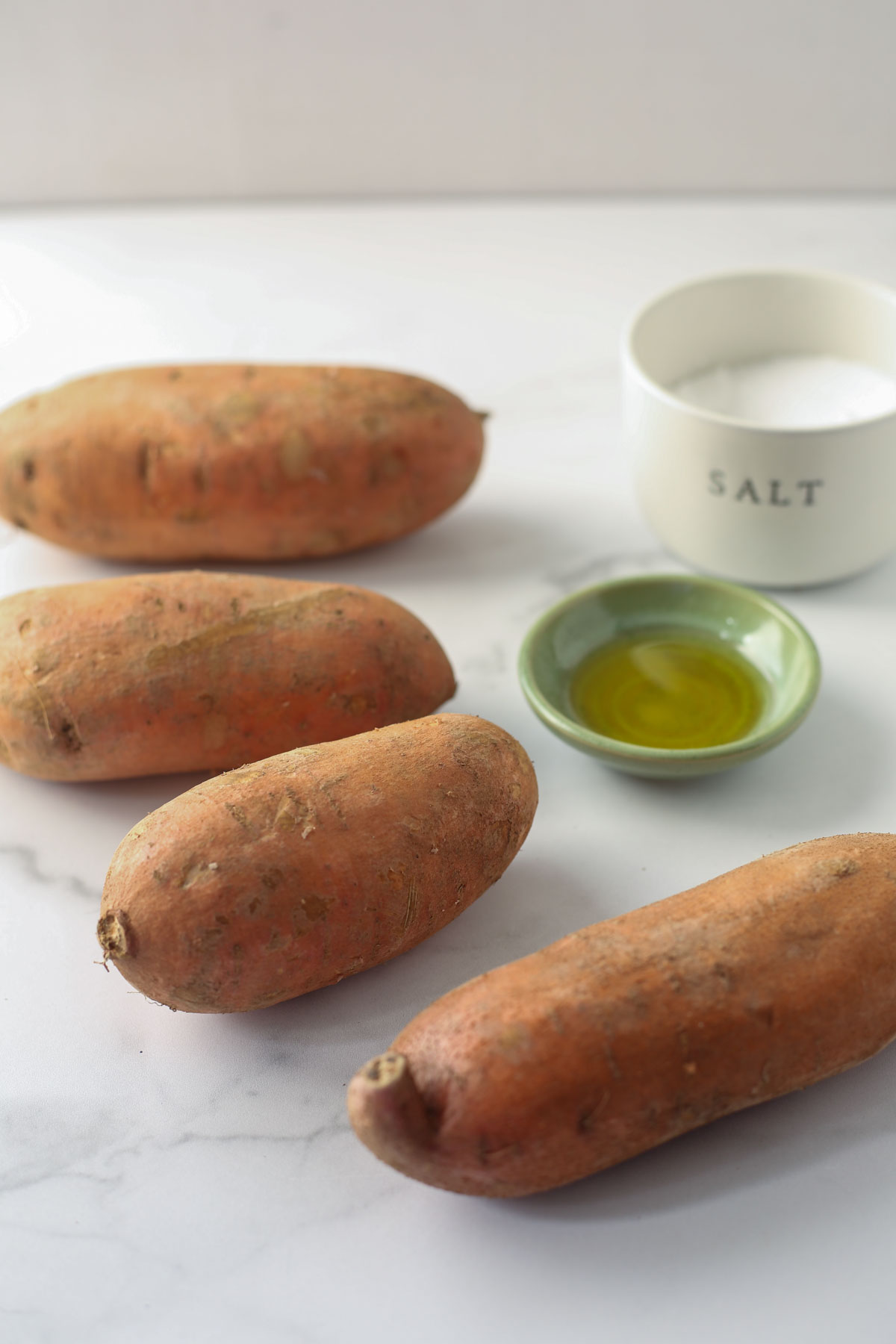 Ingredients for baked sweet potatoes lined up on a marble background. Sweet potatoes, olive oil, and salt.