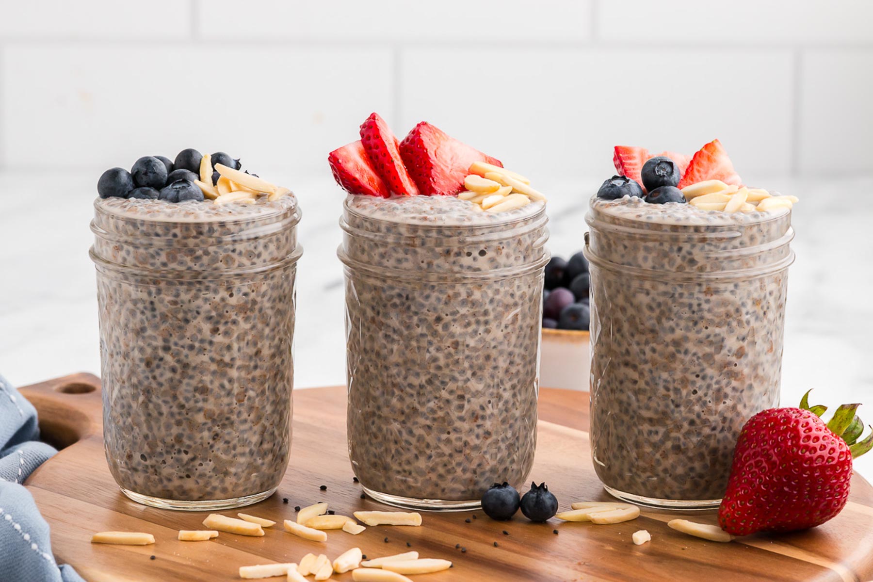 Mason jars full of basil seed pudding, three in a row, on a wooden background with almonds and berries.