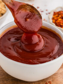 A wooden spoon drips homemade barbecue sauce back down into a white bowl. It sits on a brown wooden board with crushed red pepper flakes in the background.