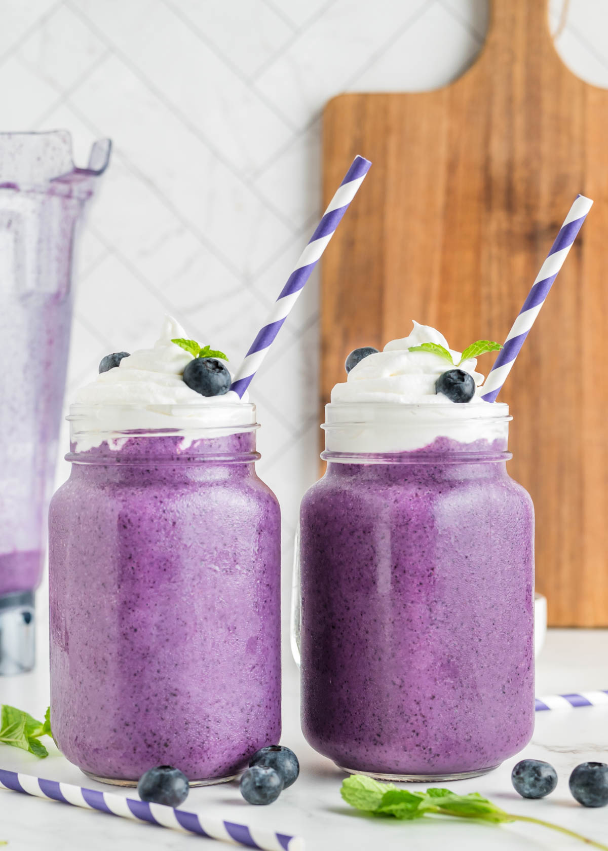 Side view of blueberry milkshakes served in a mason jar with whipped cream, blueberries, and a mint sprig. A purple and white striped straw is in the top of each glass.