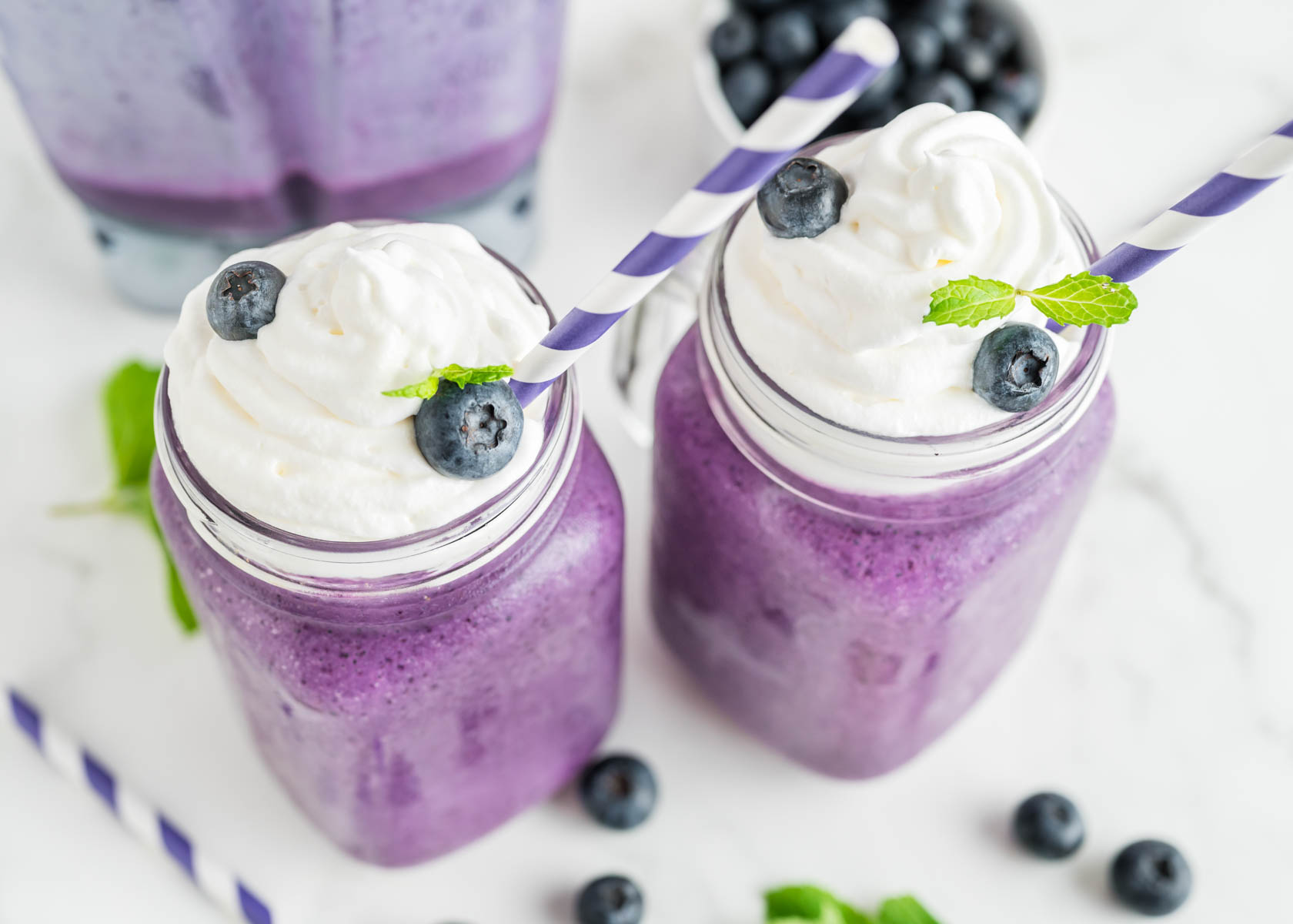 Blueberry milkshakes served in a mason jar with whipped cream, blueberries, and a mint sprig. A purple and white striped straw is in the top of each glass.