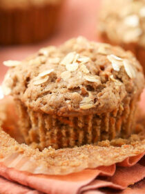 A carrot muffin sits on a rust-colored napkin with the cupcake liner peeled down.