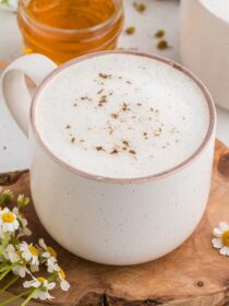 Chamomile Latte in a white mug, sitting on a wooden board and surrounded by fresh chamomile flowers