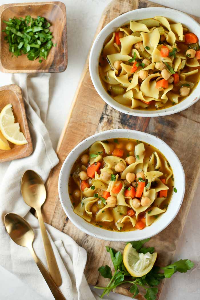 two bowls of chickpea noodle soup on a board with parsley and lemon slices on the side to use as garnish.