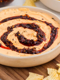 Chili Crisp Dip in a white bowl with a swirl of chili crisp on the top. The bowl sits on a wooden board with potato chips scattered around.