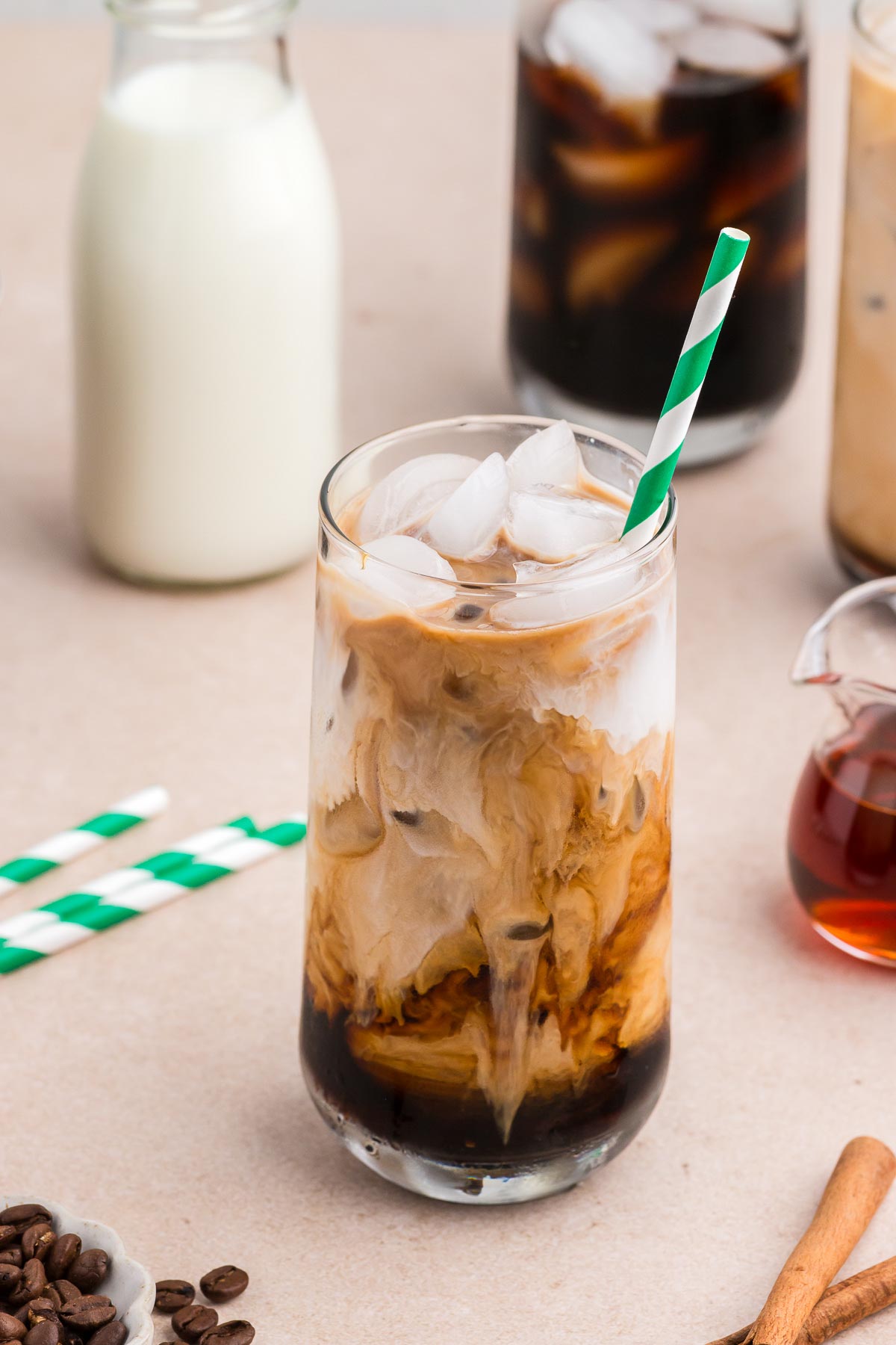 A tall clear glass of cinnamon dolce iced coffee with a green and white striped straw plus coffee, cream, and syrup in the background