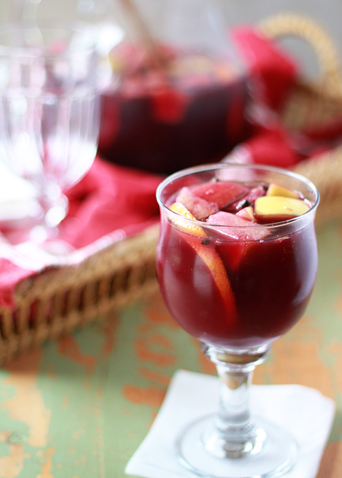 A glass of red wine sangria full of fruit and wine. It sits on a green background with a serving tray behind it. 