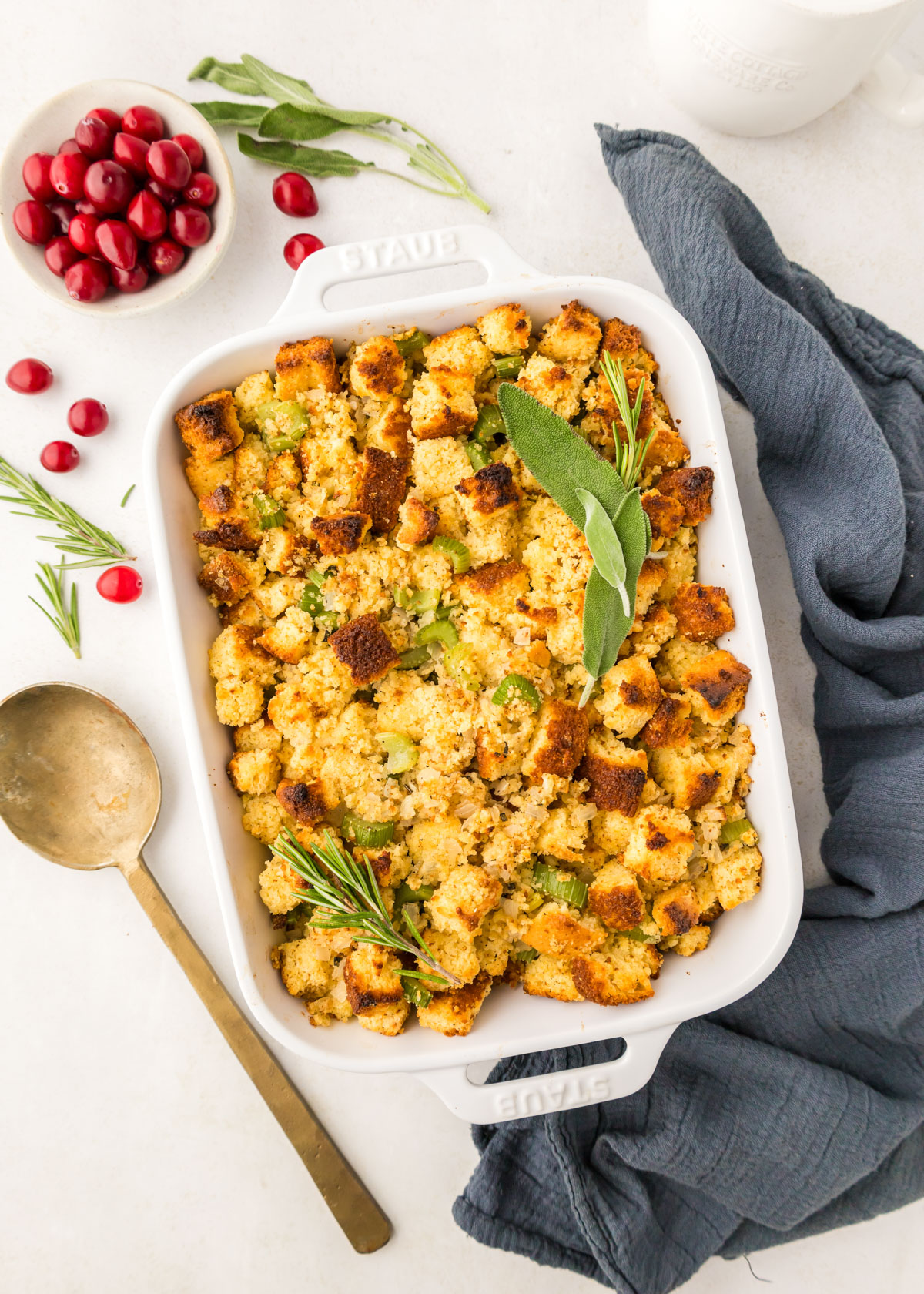 Cornbread Stuffing in a white casserole dish with a dark blue napkin, gold serving spoon, rosemary and cranberries in the background. 
