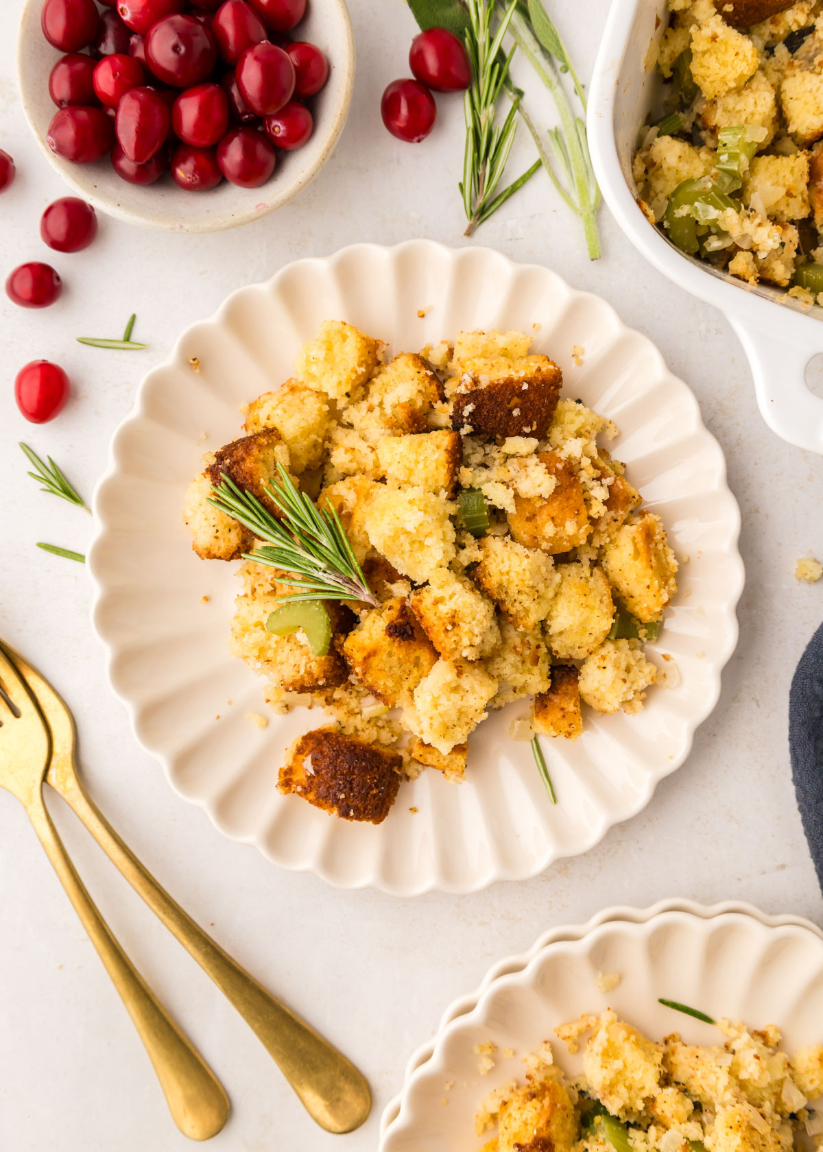 A serving of cornbread stuffing on a white scalloped plate with cranberries and rosemary in the background.