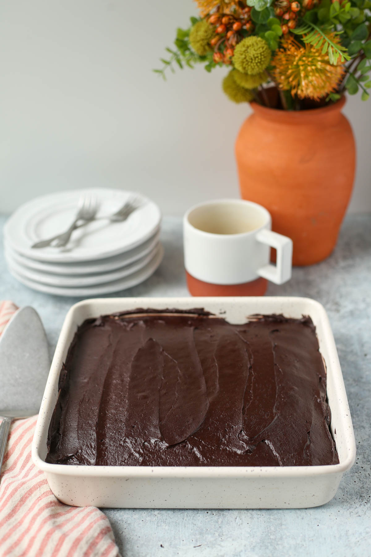 chocolate wacky cake in a white metal pan with coffee, plates, and flowers in the background.