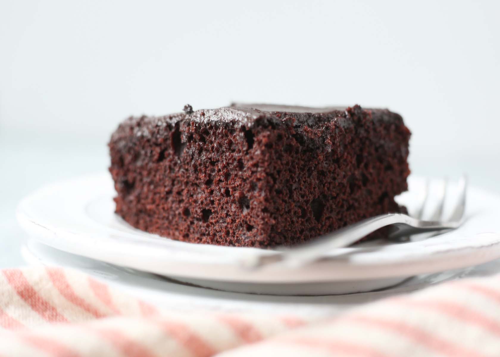 A piece of chocolate wacky cake on a white plate with a fork and a striped napkin.