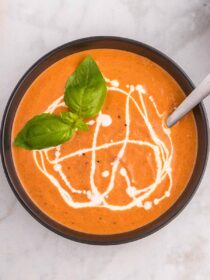 A black bowl filled with creamy tomato soup with a drizzle of heavy cream and a sprig of fresh basil.