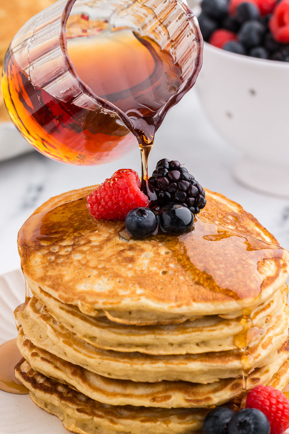a clear pitcher of pure maple syrup being drizzled onto a stack of pancakes with fruit on top