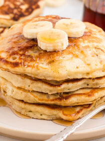 Golden fluffy pancakes in a stack with three banana slices and maple syrup running down the sides