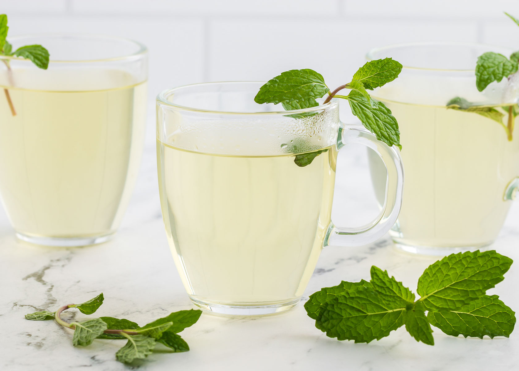 a clear glass mug of fresh mint tea with fresh mint scattered around.