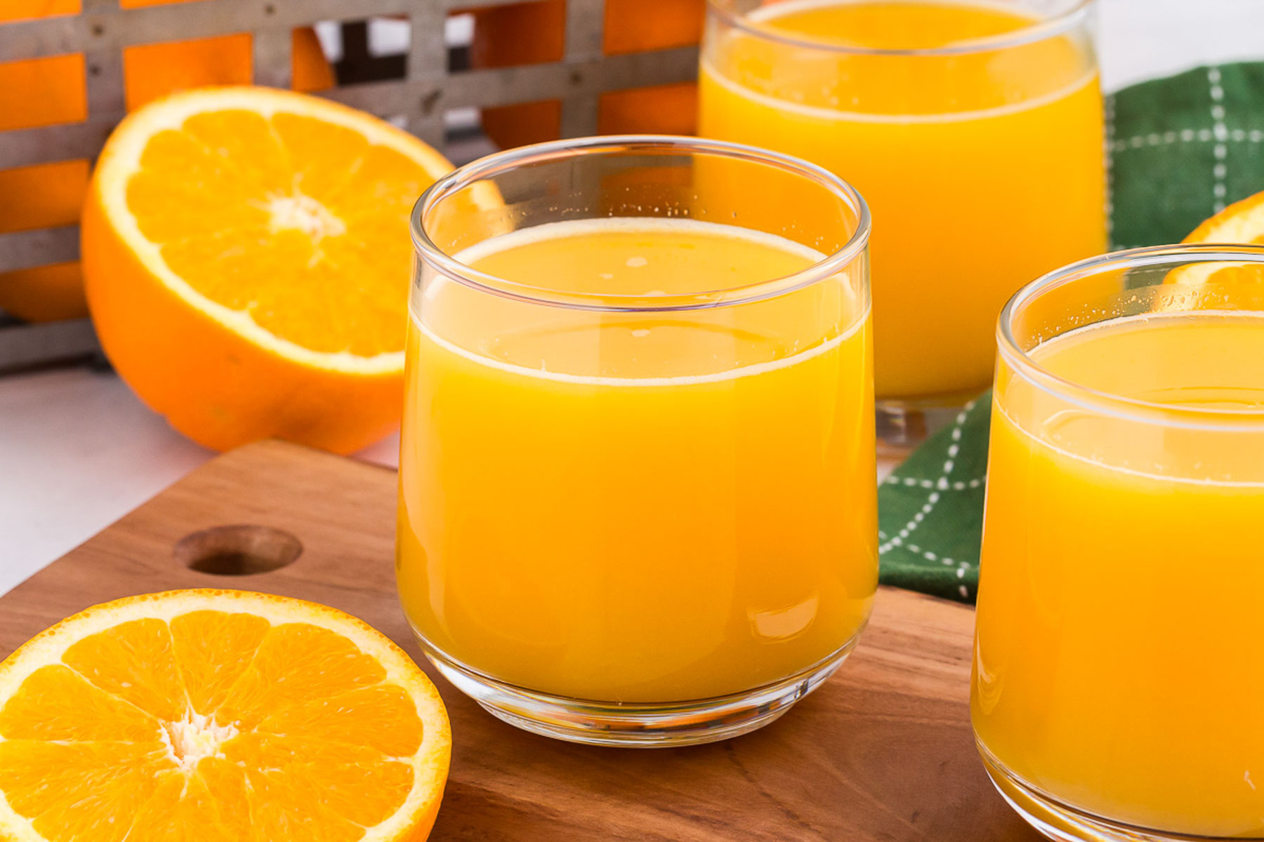 Horizontal image showing a clear glass full of orange juice on a wooden board surrounded by sliced oranges and a green napkin.