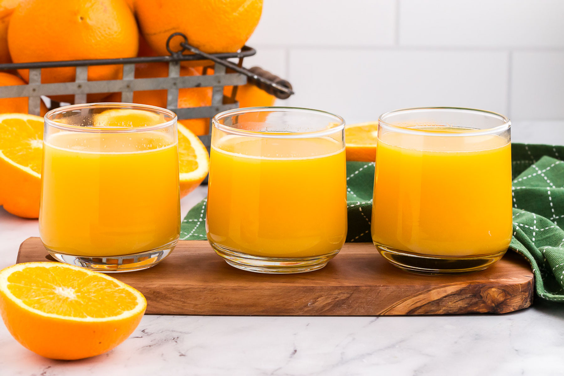 Three glasses in a row of fresh squeezed orange juice on a wooden board with a green towel in the background.