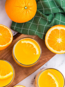 A top view of two glasses of orange juice with fresh orange slices for garnish. A green towel is in the background.