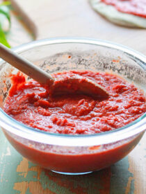 A bowl of pizza sauce made with fresh tomatoes. The bowl is clear glass, with a wooden spoon in it. The background is sage green distressed wood.