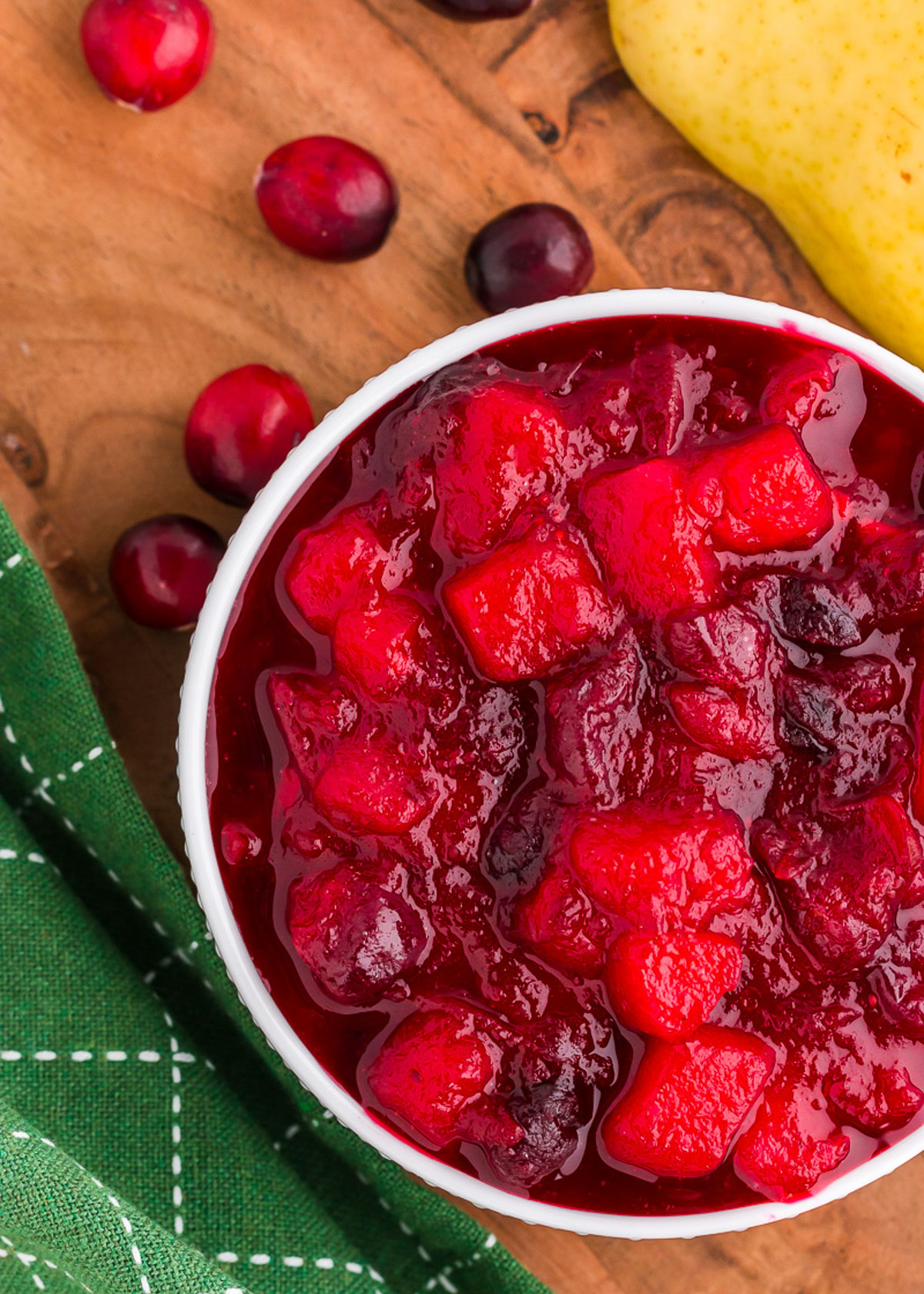 A top view of ginger pear cranberry sauce in a white bowl on a wood table with a green napkin in the background.