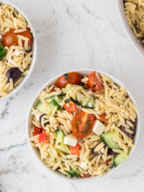 Two white bowls full of greek orzo pasta salad with a serving bowl. They sit on a marble background.