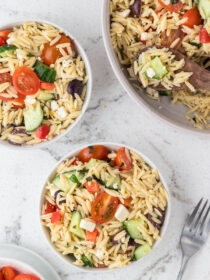 Two white bowls full of greek orzo pasta salad with a serving bowl. They sit on a marble background.