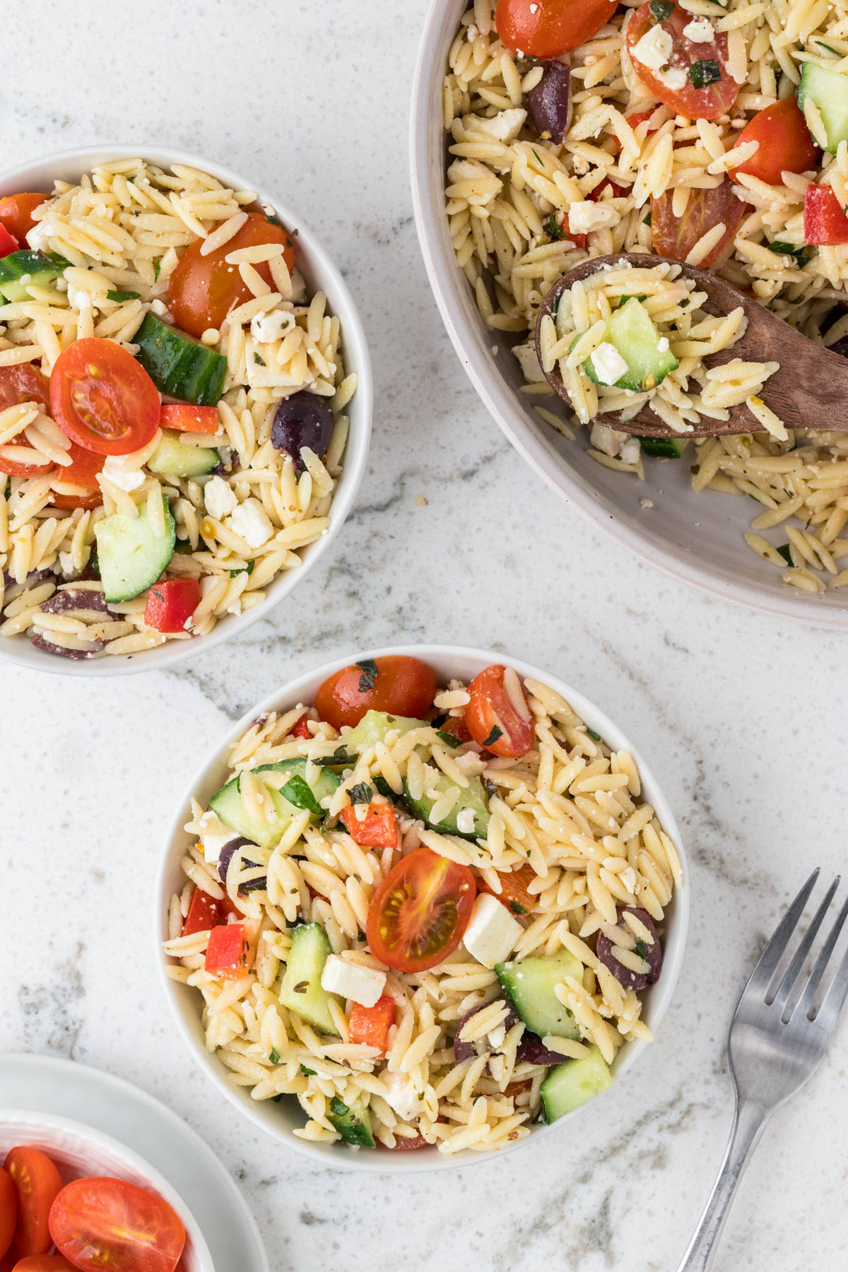 Two white bowls full of greek orzo pasta salad with a serving bowl. They sit on a marble background.
