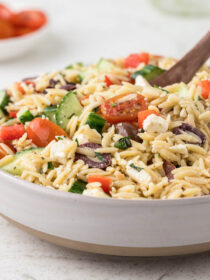 A white bowl full of greek orzo pasta salad with cherry tomatoes in the background.