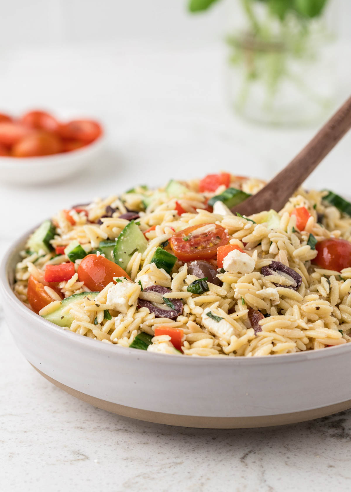 A white bowl full of greek orzo pasta salad with cherry tomatoes in the background.