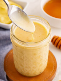 A jar of honey dijon mustard dressing on a brown coaster with a blue napkin in the background. A spoon is scooping some of the dressing out.