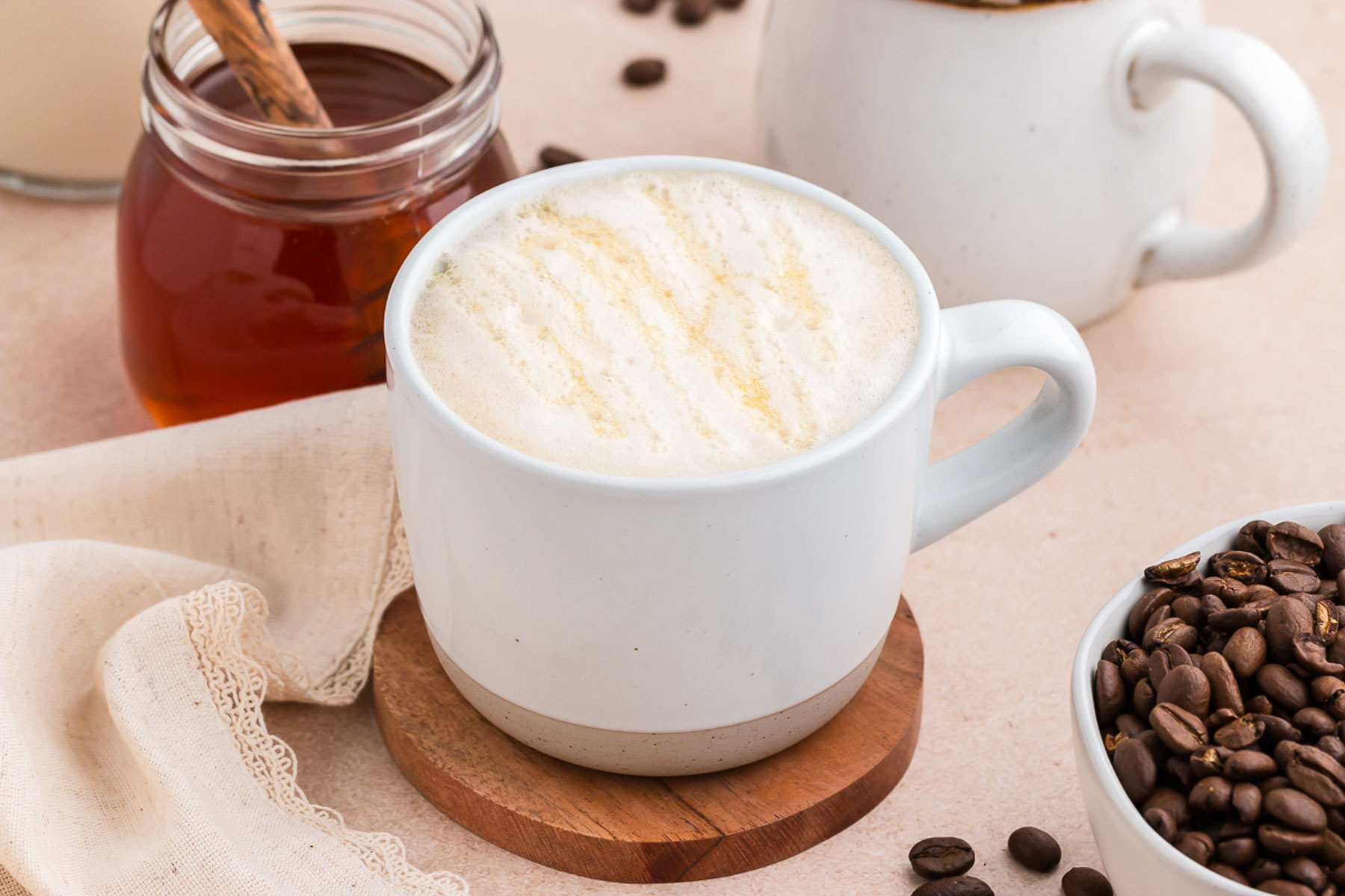 A white mug full of oat milk latte with coffee beans and honey in the background