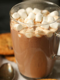 Hot chocolate in a clear mug with marshmallows on top and chocolate chip cookies.