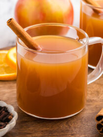 Two mugs of hot spiced mulled cider in clear mugs with a cinnamon stick in as garnish. Sliced orange, cloves, and apples are in the background.