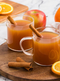 Two mugs of hot spiced mulled cider in clear mugs with a cinnamon stick in as garnish. Sliced orange, cloves, and cinnamon sticks are in the background.
