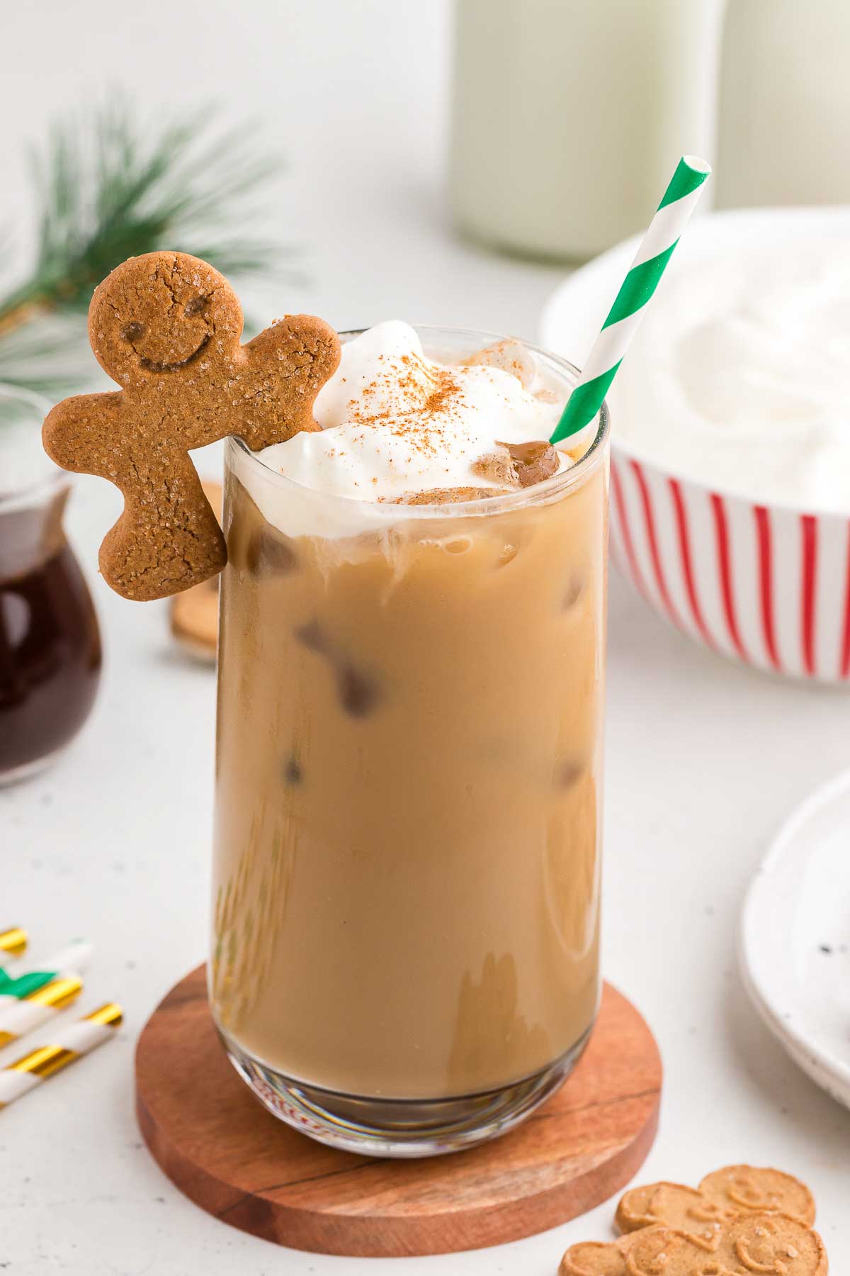 Gingerbread Iced Latte in a clear glass with a green and white striped straw, whipped cream, and a gingerbread cookie perched on the rim of the glass. 