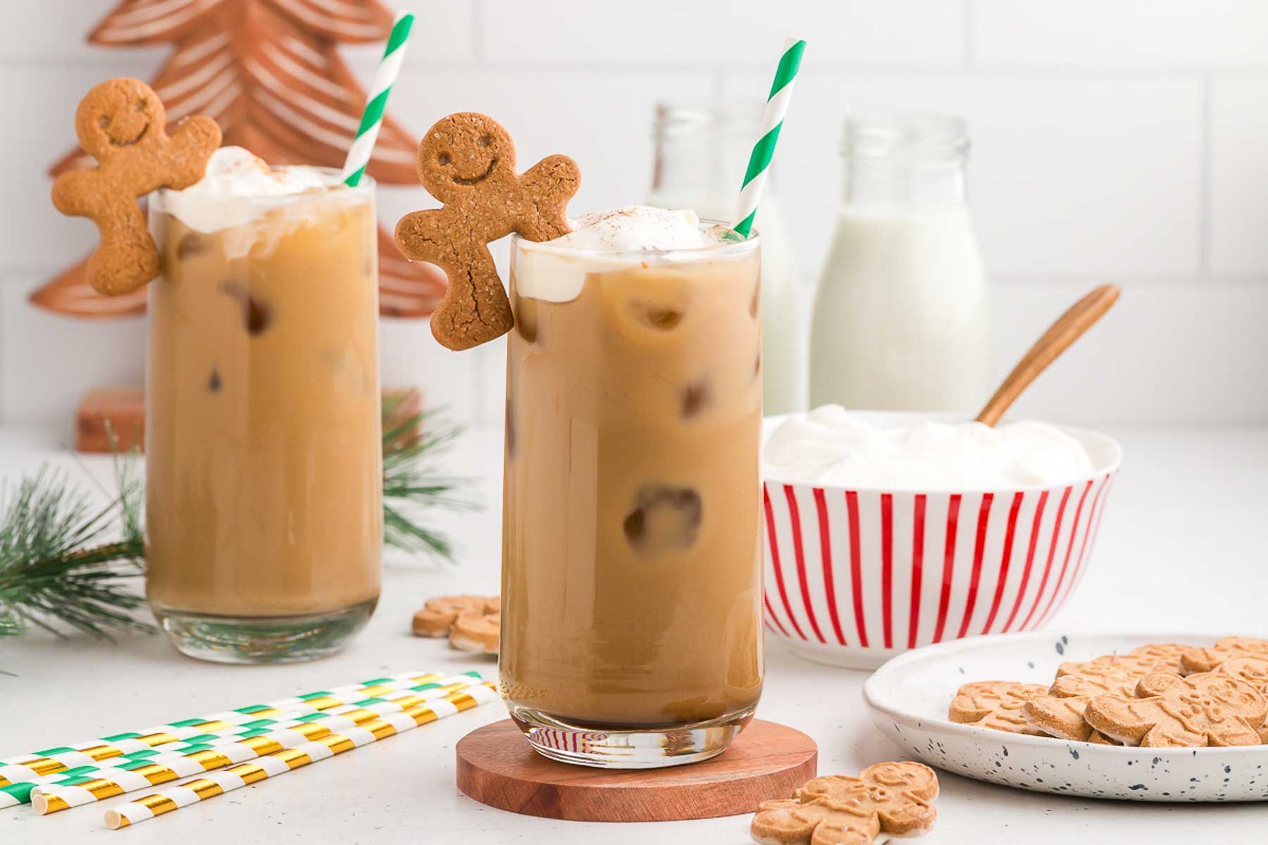Two Gingerbread Iced Lattes with green and white striped straws and gingerbread cookies perched on top. A bowl of whipped cream in a red and white striped bowl is in the background. 