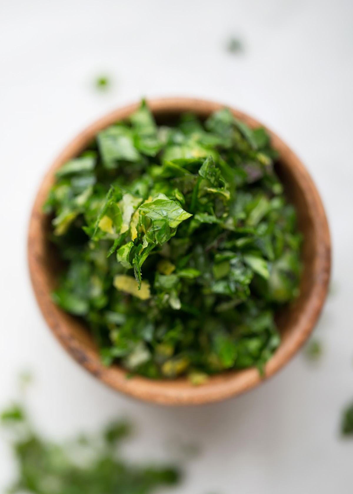 Small wooden bowl full of mint gremolata on a white plaster background, with some more gremolata in the foreground. 