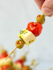 A hand holds a marinated cheese and olive skewer.