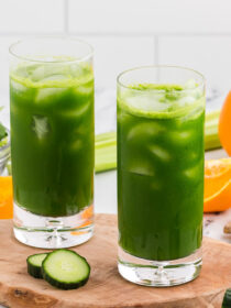 Two glasses of green juice on a wooden board with veggies and fruit in the background.