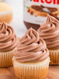 Three vanilla cupcakes topped with fluffy Nutella buttercream frosting and a jar of Nutella in the background.