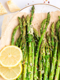 A top view of a white plate lined with parchment paper and parmesan roasted asparagus on top with lemon as garnish.