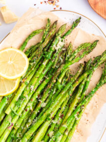 A top view of a white plate lined with parchment paper and parmesan roasted asparagus on top with lemon as garnish.