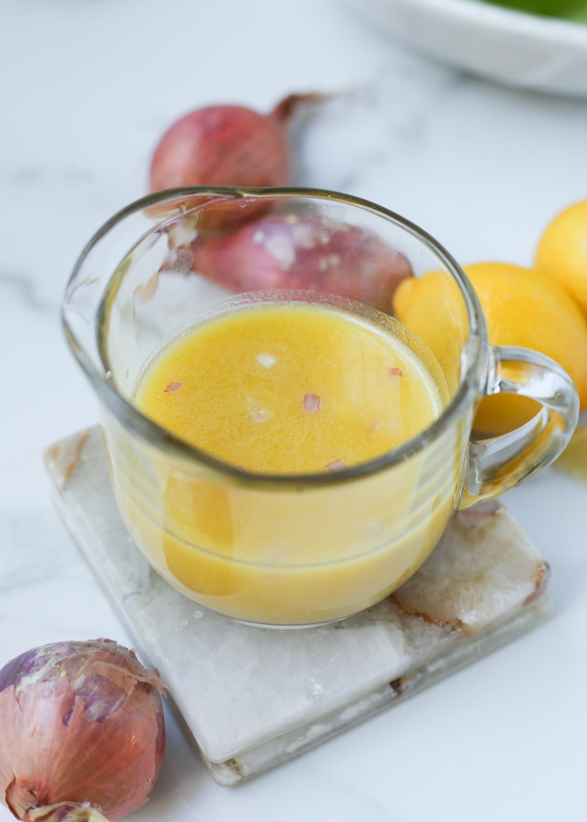 A pitcher full of shallot vinaigrette with shallots and lemons in the background.