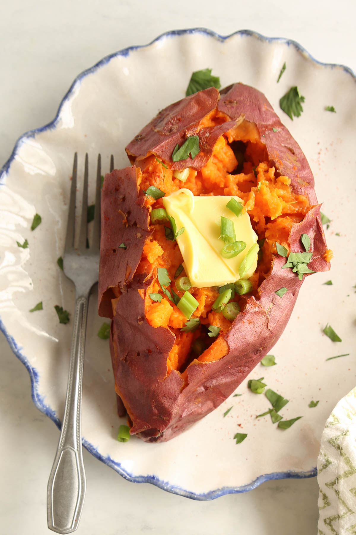 A top view of a baked sweet potato with a pat of butter on an organic shaped white and blue plate and with a silver fork.