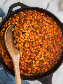 A wooden spoon scoops vegetarian baked beans (from canned beans) out of a skillet