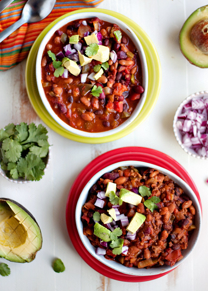 A bowl of 3-bean chili in a white bowl on a lime green plate with avocados, red onion, and cilantro on top along with another version with meat added in a white bowl on a red plate. 